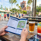 Person holding a park map with a drink and smartphone on a table, surrounded by palm trees and people.