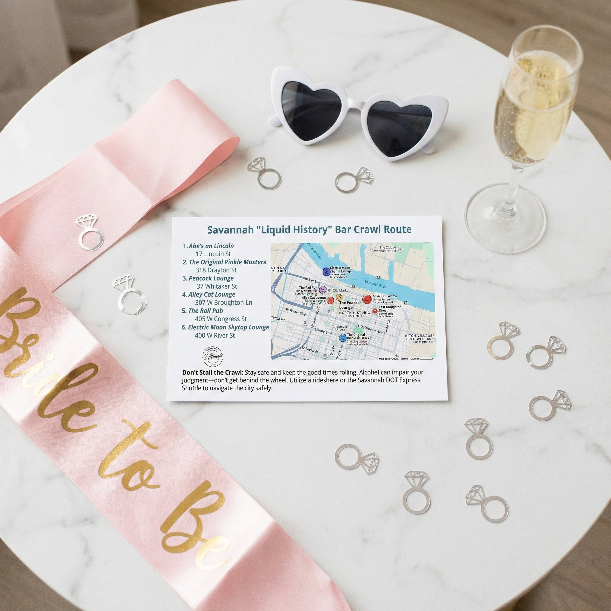 Bachelorette party items including a pink sash, sunglasses, map, and champagne on a marble table.